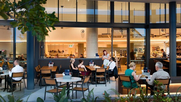 Un área de comedor al aire libre con mesas y sillas, una zona de bar al fondo y vistas del interior a través de amplios ventanales.