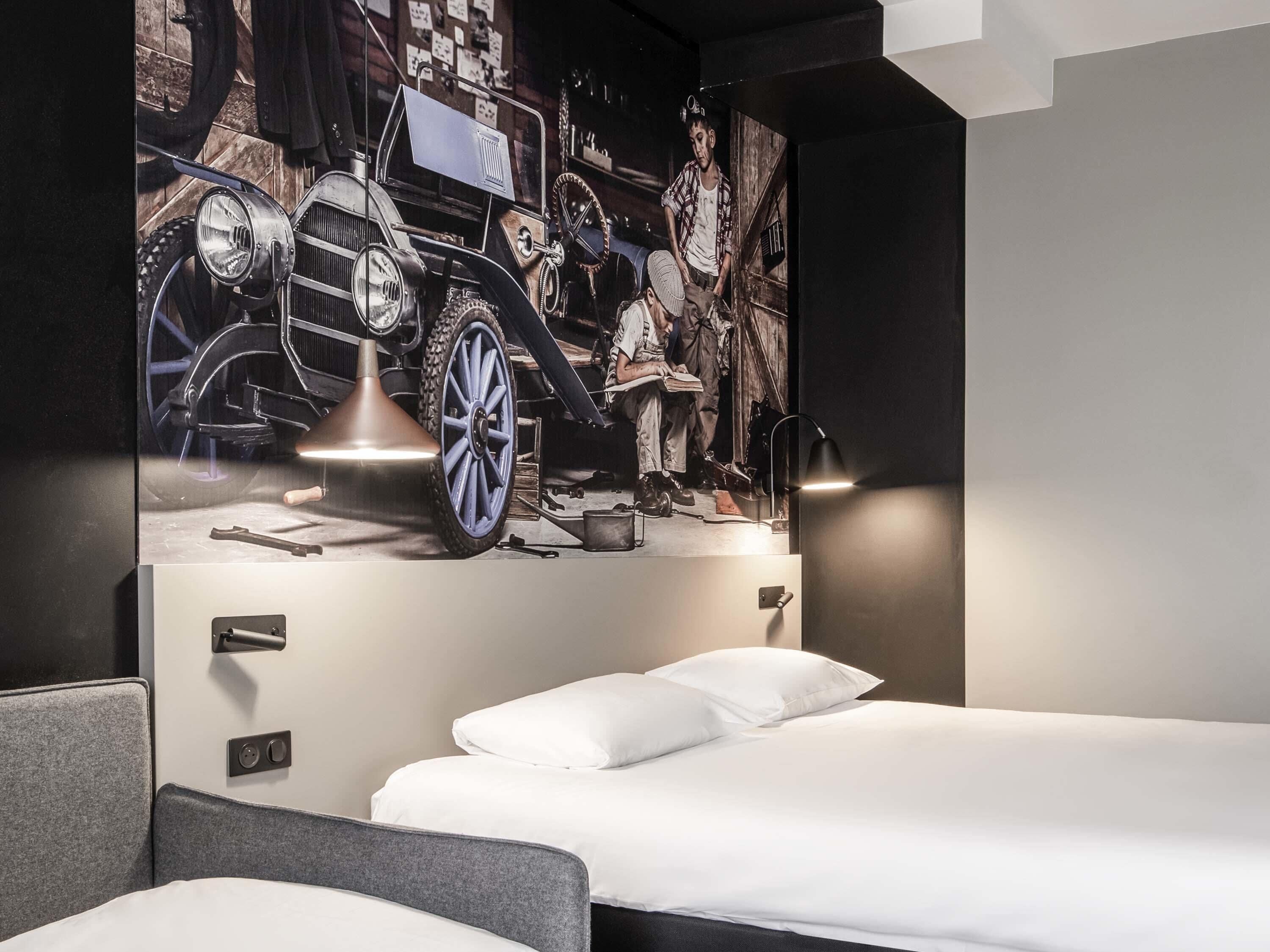 Una habitación de hotel moderna con un gran mural de coches antiguos, una cama con ropa de cama blanca y una lámpara de escritorio.