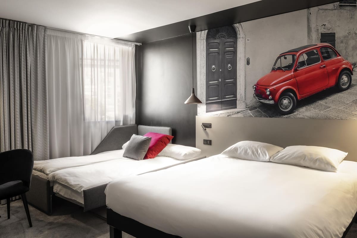 Een hotelkamer met twee bedden, een stoel, een raam met gordijnen, een wandlamp en een grote poster van een rode auto.