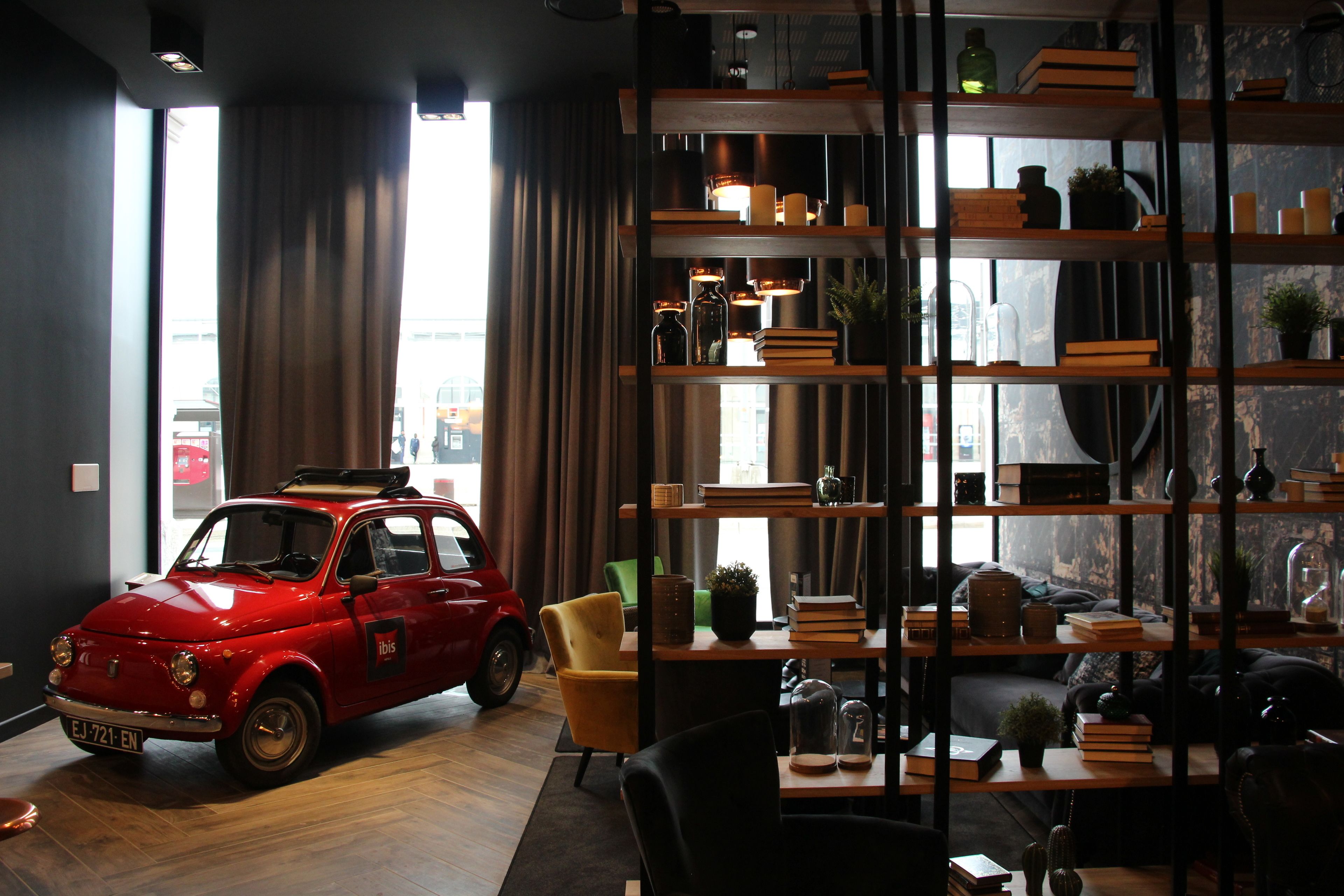 A room with a vintage red car, a wooden bookshelf, and a view of the outside through large windows.
