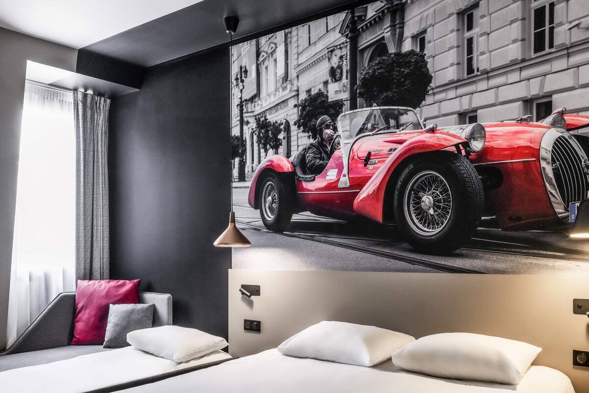 Een moderne hotelkamer met een groot muurschildering van een klassieke rode auto, een bed met witte lakens, en een grijze bank met rode en grijze kussens.