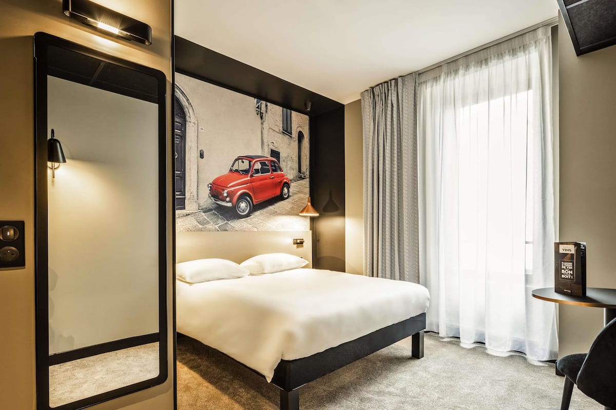 Hotelkamer met een bed, een bureau, een stoel, een lamp, een schilderij van een auto en een raam met gordijnen.
