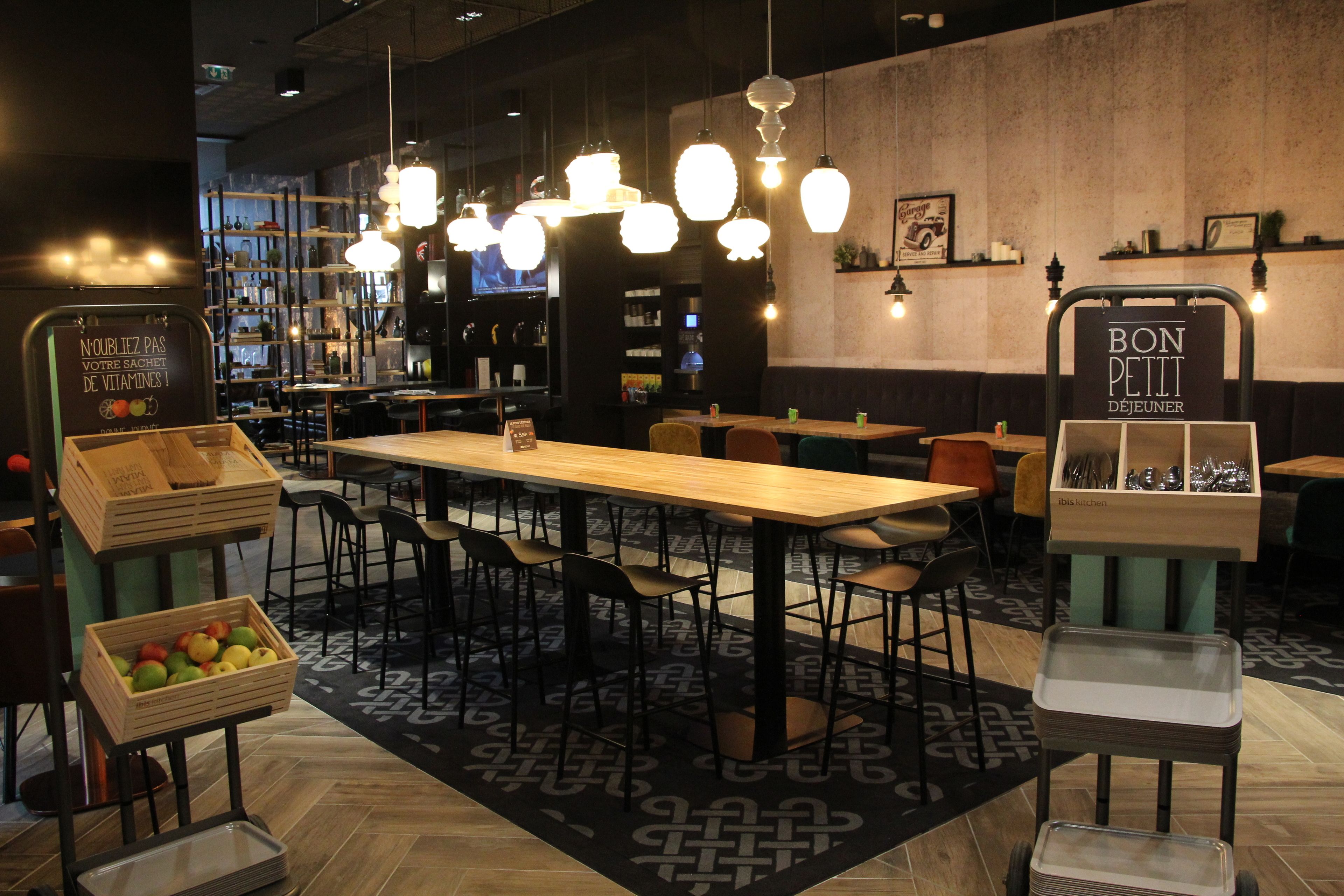 L’intérieur d’un restaurant avec des tables en bois, des chaises noires, des suspensions et un tapis à motifs.