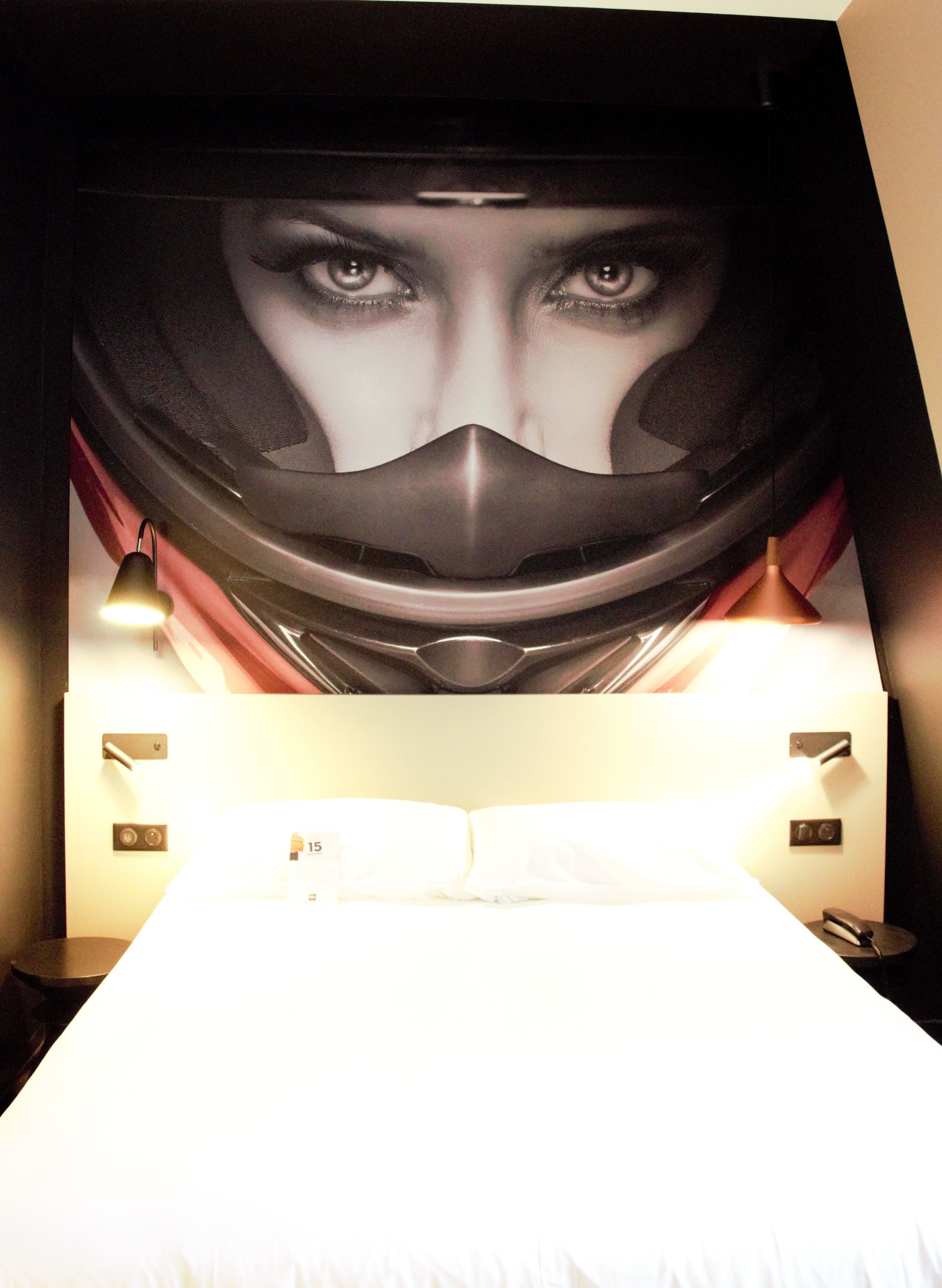 Quarto de hotel com um pôster grande na parede, uma cama, duas luminárias de cabeceira e uma mesa de cabeceira com um telefone.