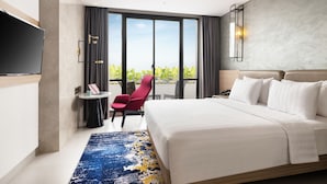 Ein modernes Hotelzimmer mit einem großen Bett, einem Fernseher, einem kleinen Tisch mit Stuhl und Blick auf Grün hinter großen Fenstern.