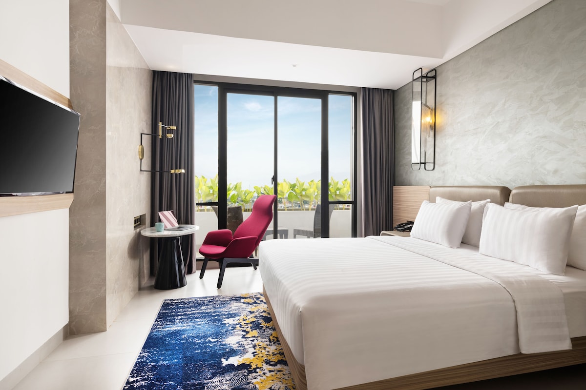 Une chambre d’hôtel moderne dotée d’un grand lit, d’une télévision, d’une petite table avec une chaise et offrant une vue sur la verdure grâce à de grandes fenêtres.