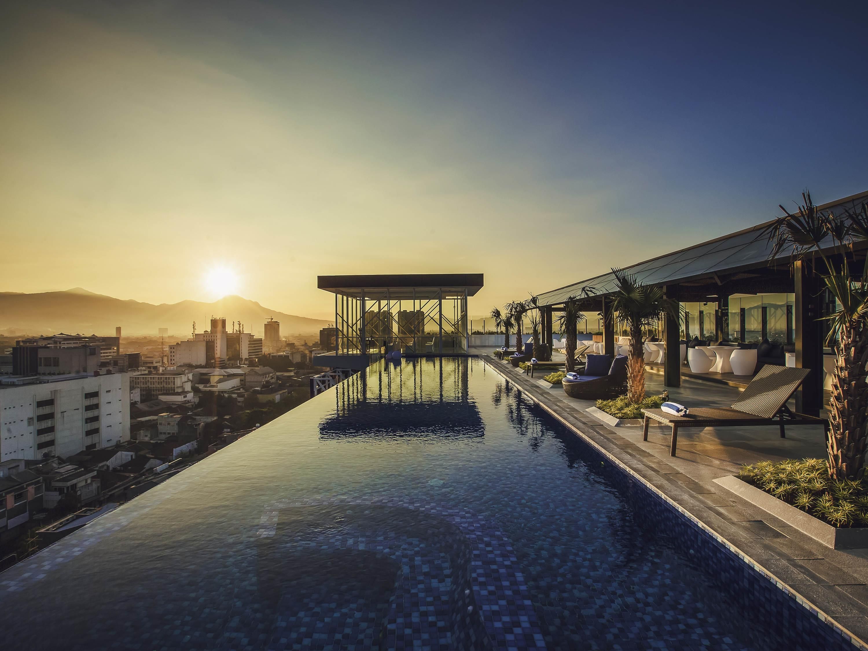 Piscina infinita no terraço com vista para o horizonte urbano, cadeiras de descanso e área de estar coberta.