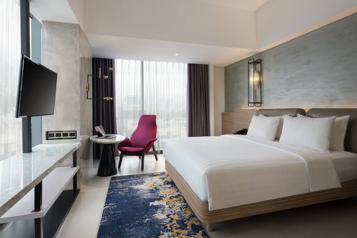 Ein modernes Hotelzimmer mit einem großen Bett, einem Schreibtisch mit Flachbildfernseher, einem Sessel und Blick auf die Stadt durch große Fenster.