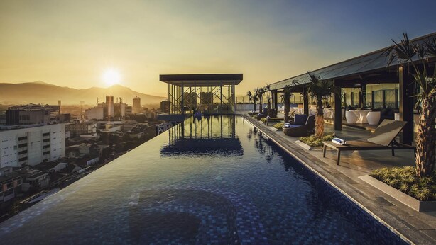 Ein Infinity-Pool auf dem Dach mit Blick auf die Stadtlandschaft, Liegestühle und eine überdachte Sitzgelegenheit.