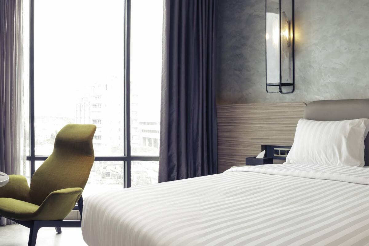 Une chambre d’hôtel avec un grand lit, une chaise, une table de chevet et une vue sur la ville grâce à de grandes fenêtres.