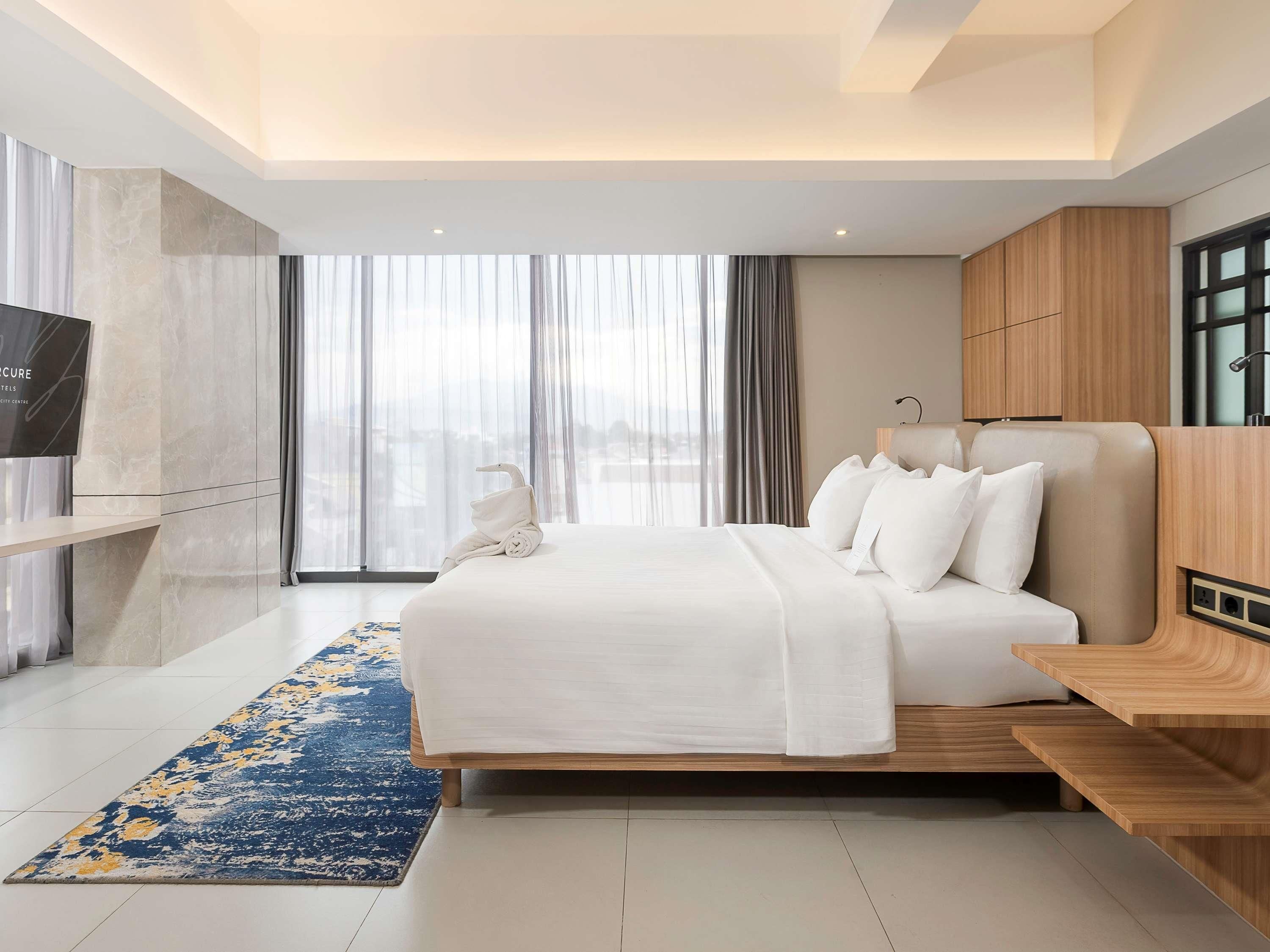 A modern hotel room with a large bed, a TV, a wooden nightstand, and a view of the city through large windows.