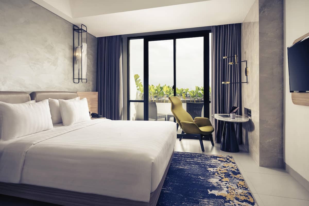Une chambre d’hôtel moderne avec un grand lit, une petite table ronde, une chaise et une grande fenêtre donnant sur un paysage verdoyant.