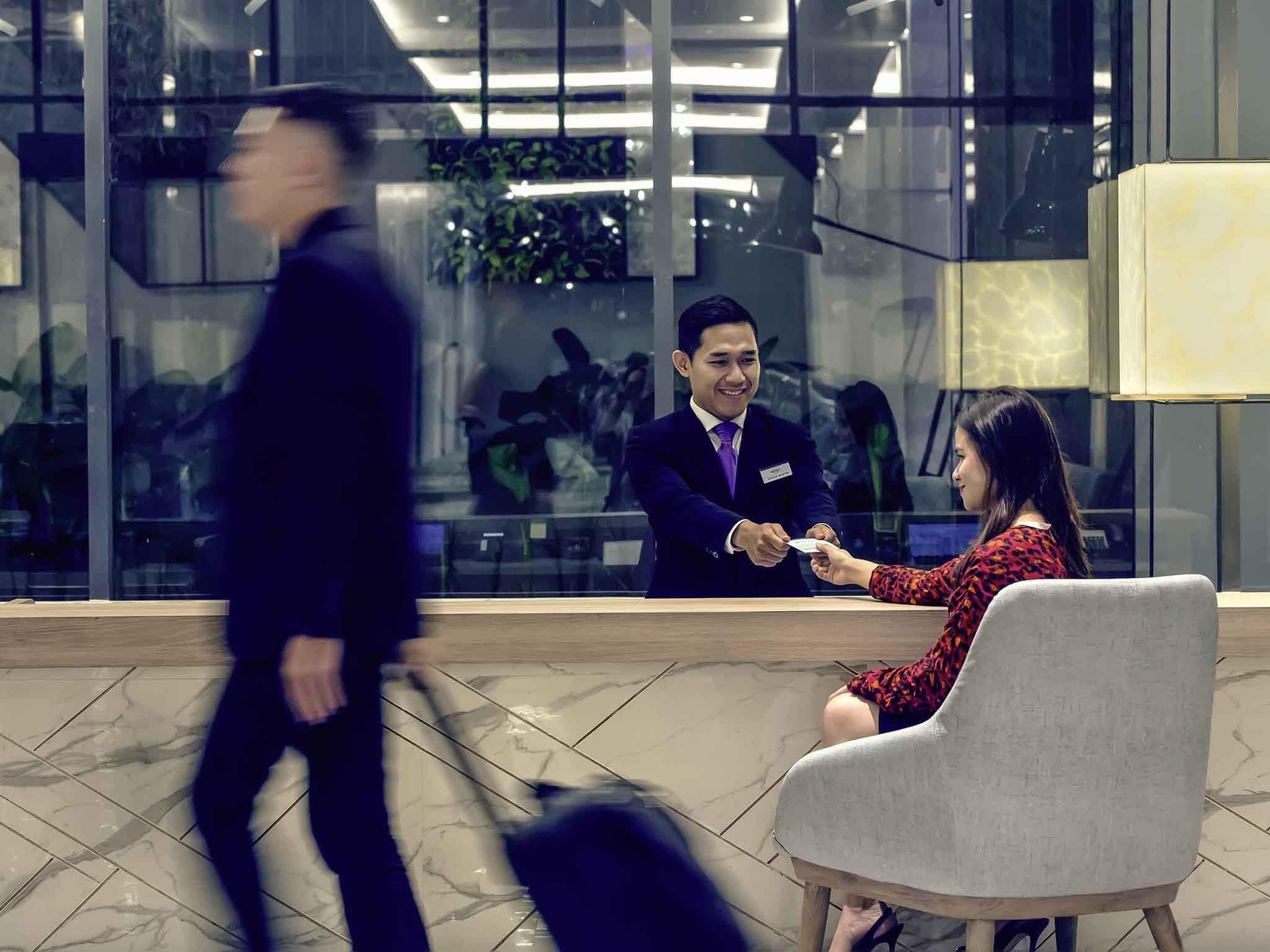 Um saguão com uma mesa de recepção, uma mulher sentada vestindo um vestido vermelho, e um homem de terno.