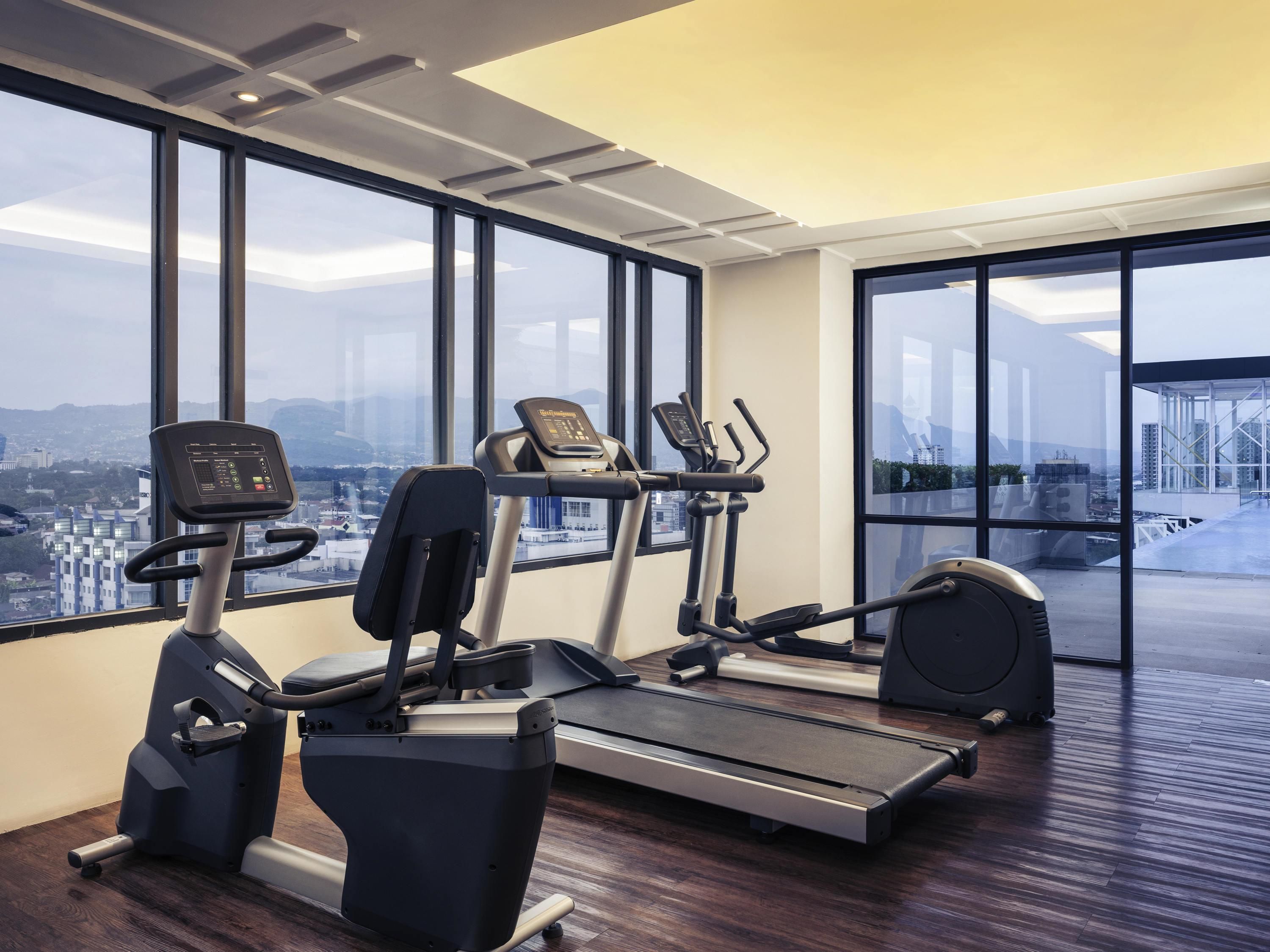 Un gimnasio con cintas de correr, elípticas y una bicicleta estática, amplias ventanas con vistas a la ciudad y suelo de madera.