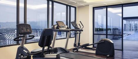 A gym with treadmills, elliptical machines, and a stationary bike, large windows with a city view, and a wooden floor.