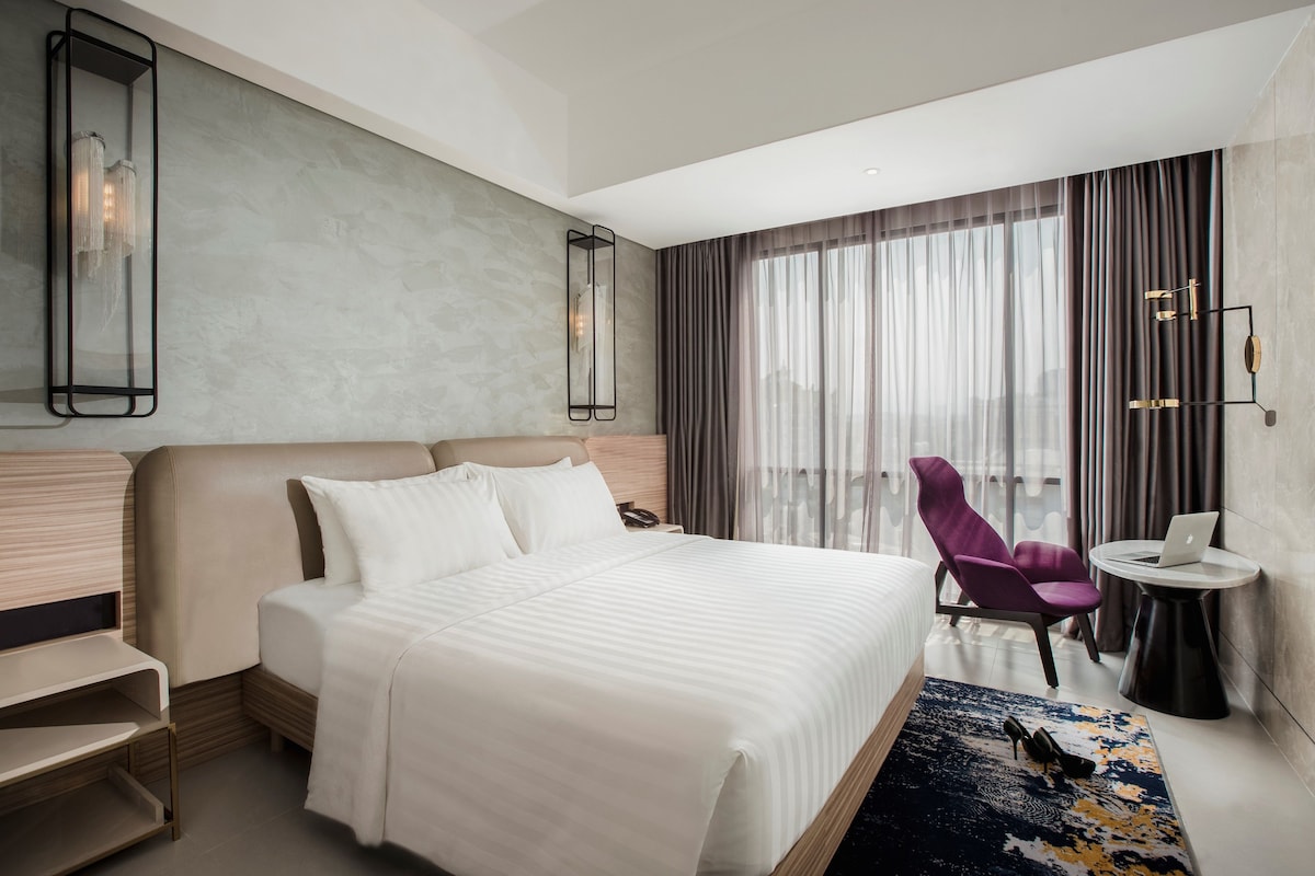 Une chambre d’hôtel moderne avec un grand lit, un bureau avec un ordinateur portable, une chaise et une vue sur la ville à travers de fines rideaux.
