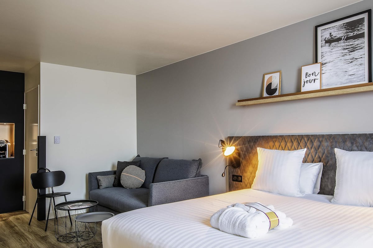 Une chambre d’hôtel moderne avec un grand lit, un canapé, une petite table et une étagère fixée au mur, ornée d’un tableau encadré et d’une pancarte.