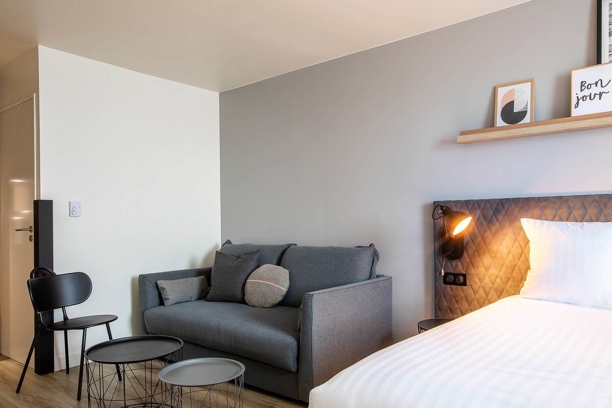 A modern hotel room with a grey sofa, a small round table, a bed with a headboard, and a wall-mounted shelf with two framed pictures.