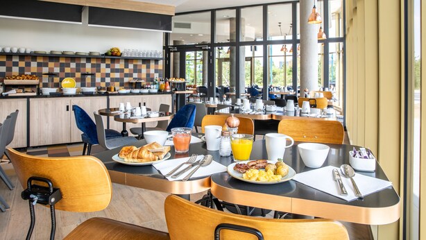 Un espace moderne dédié au petit-déjeuner, avec des tables, des chaises et une cuisine en arrière-plan.