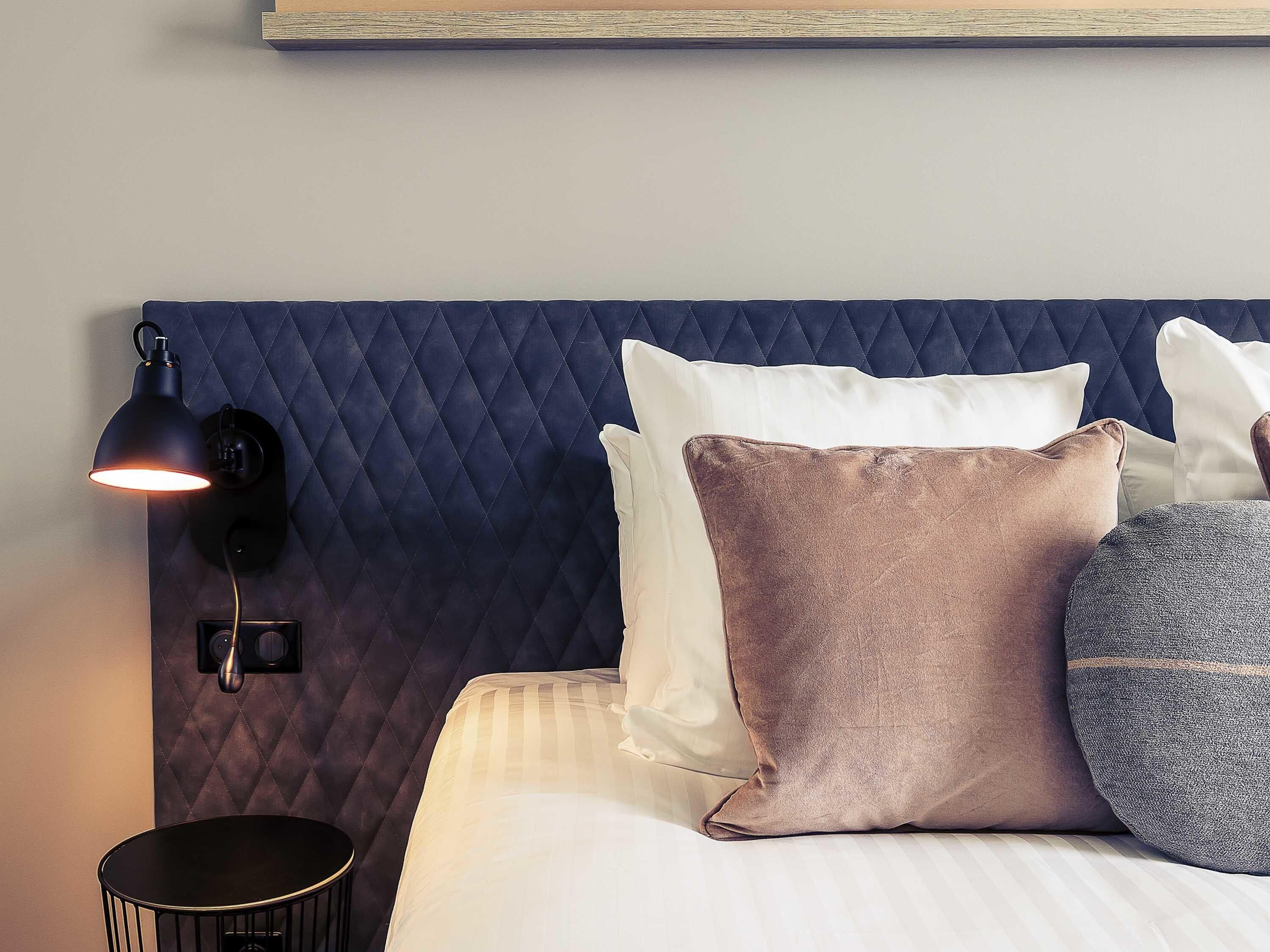 Una cama con cabecera acolchada, lámpara de noche y varias almohadas.