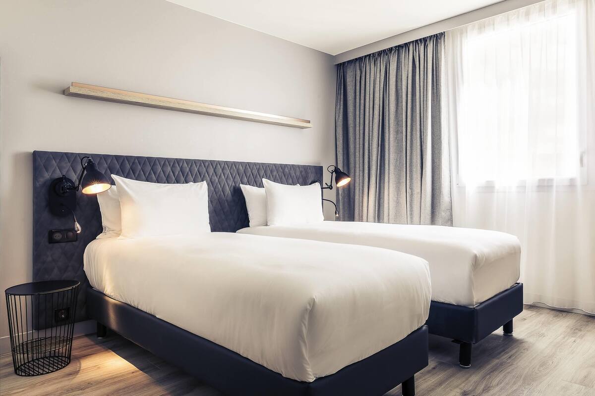Une chambre d’hôtel moderne avec deux lits, une tête de lit dotée d’un éclairage intégré, une table de chevet avec une lampe et une vue sur l’extérieur à travers des rideaux transparents.
