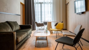 Een moderne woonkamer met een bank, een salontafel en twee stoelen. Er is een televisie aan de muur bevestigd en een raam met gordijnen.