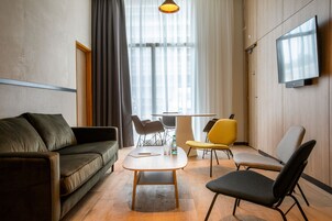 Una sala de estar moderna con un sofá, una mesa de centro y dos sillones. Hay un televisor montado en la pared y una ventana con cortinas.