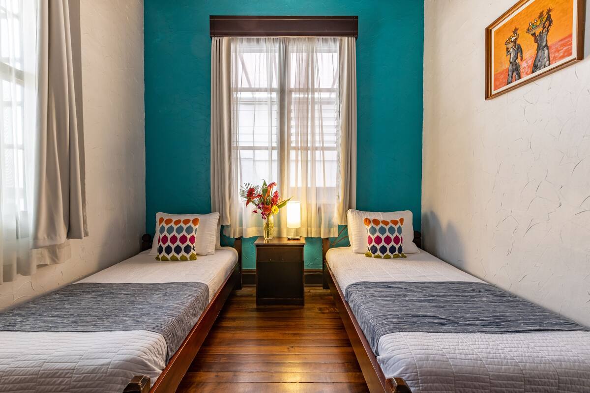 Una habitación con dos camas, una mesita con lámpara y un ventanal con cortinas.