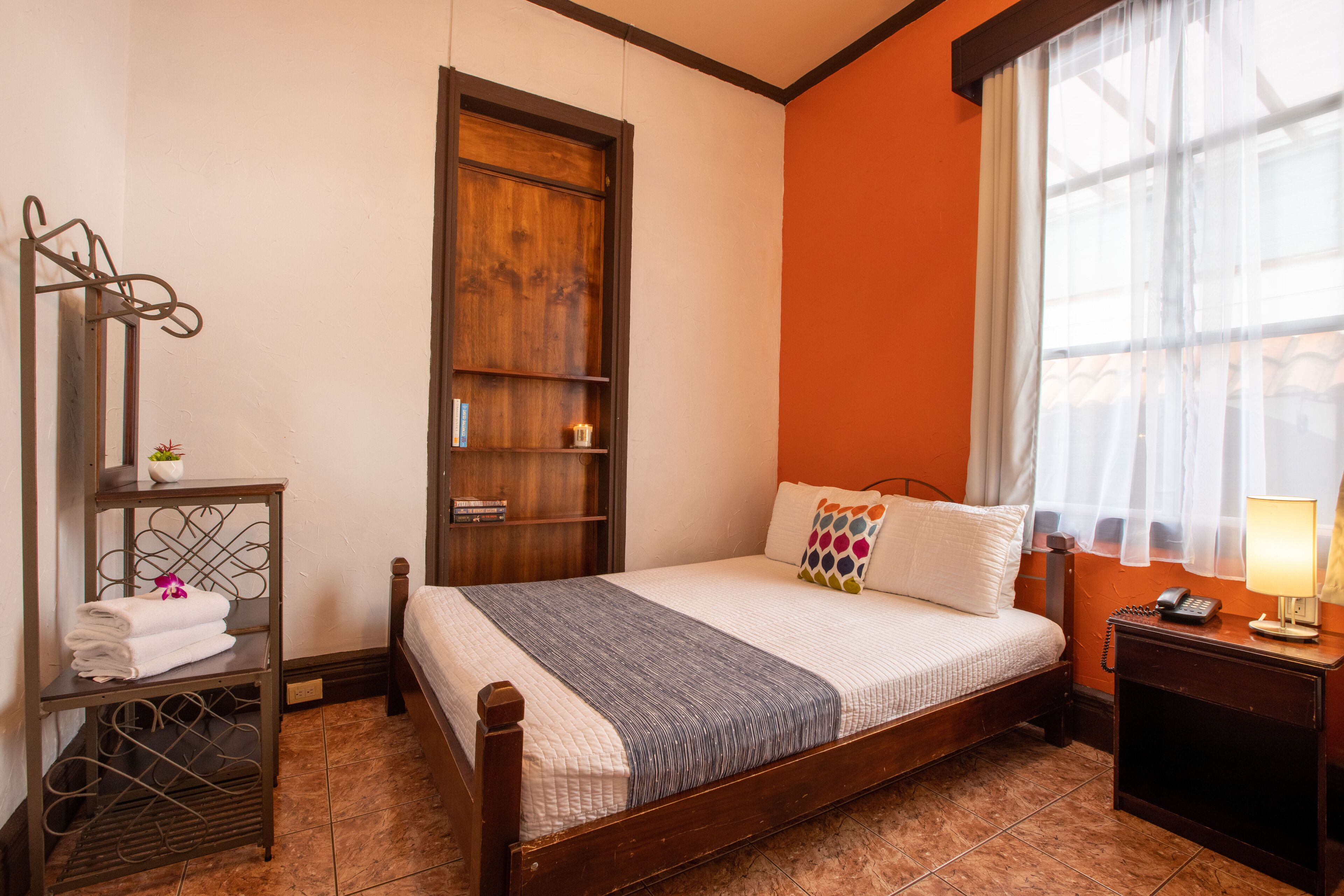 Una cama bien tendida con estructura de madera, una mesita de noche con lámpara, un estante pequeño con toallas y una ventana con cortinas translúcidas.