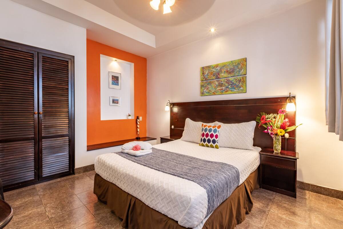Habitación de hotel con una cama grande, cabecera de madera, mesita de noche con un florero y un cuadro en la pared.