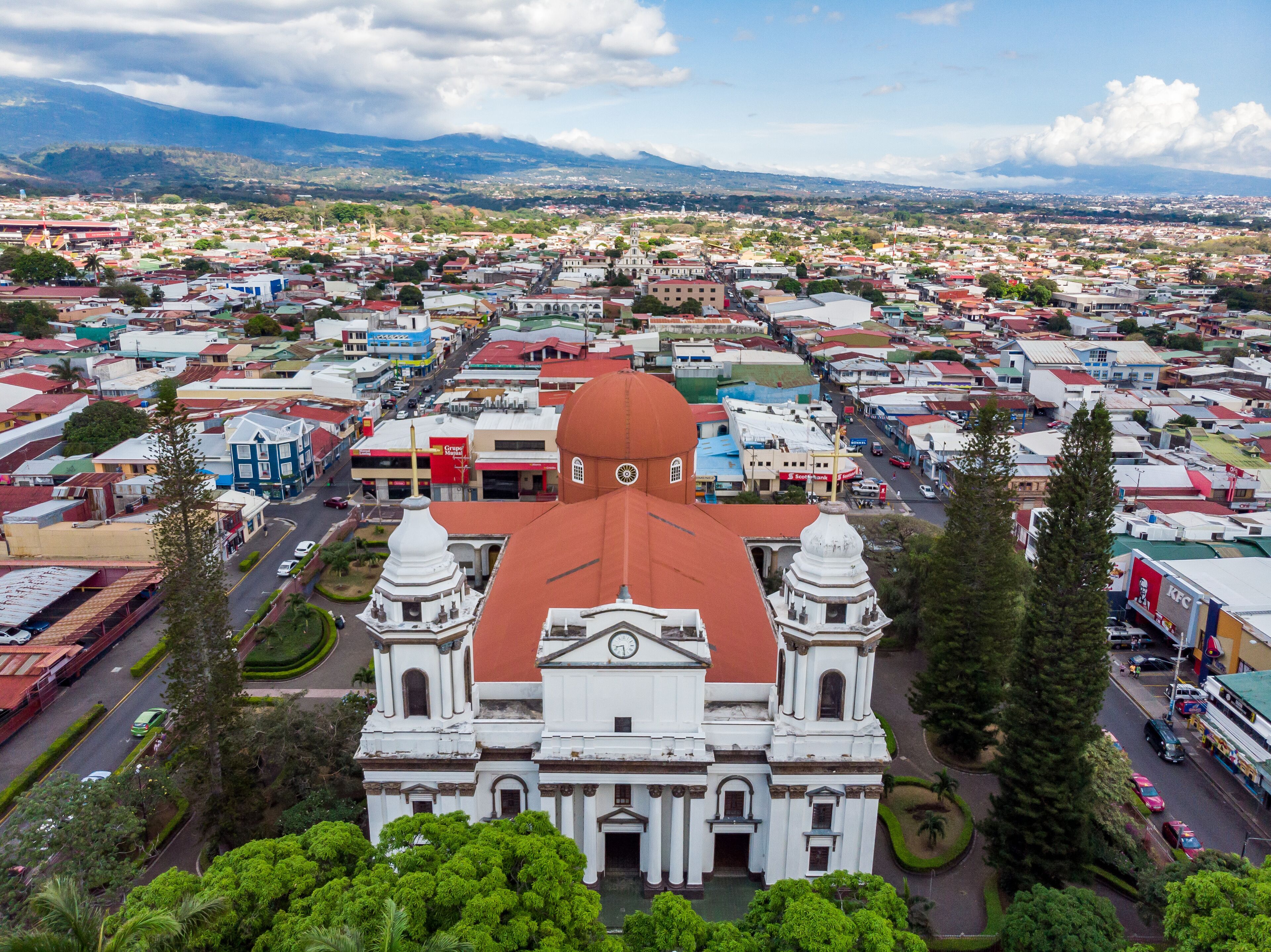 A large white building with a red roof and two domes, surrounded by a cityscape with various buildings and greenery.