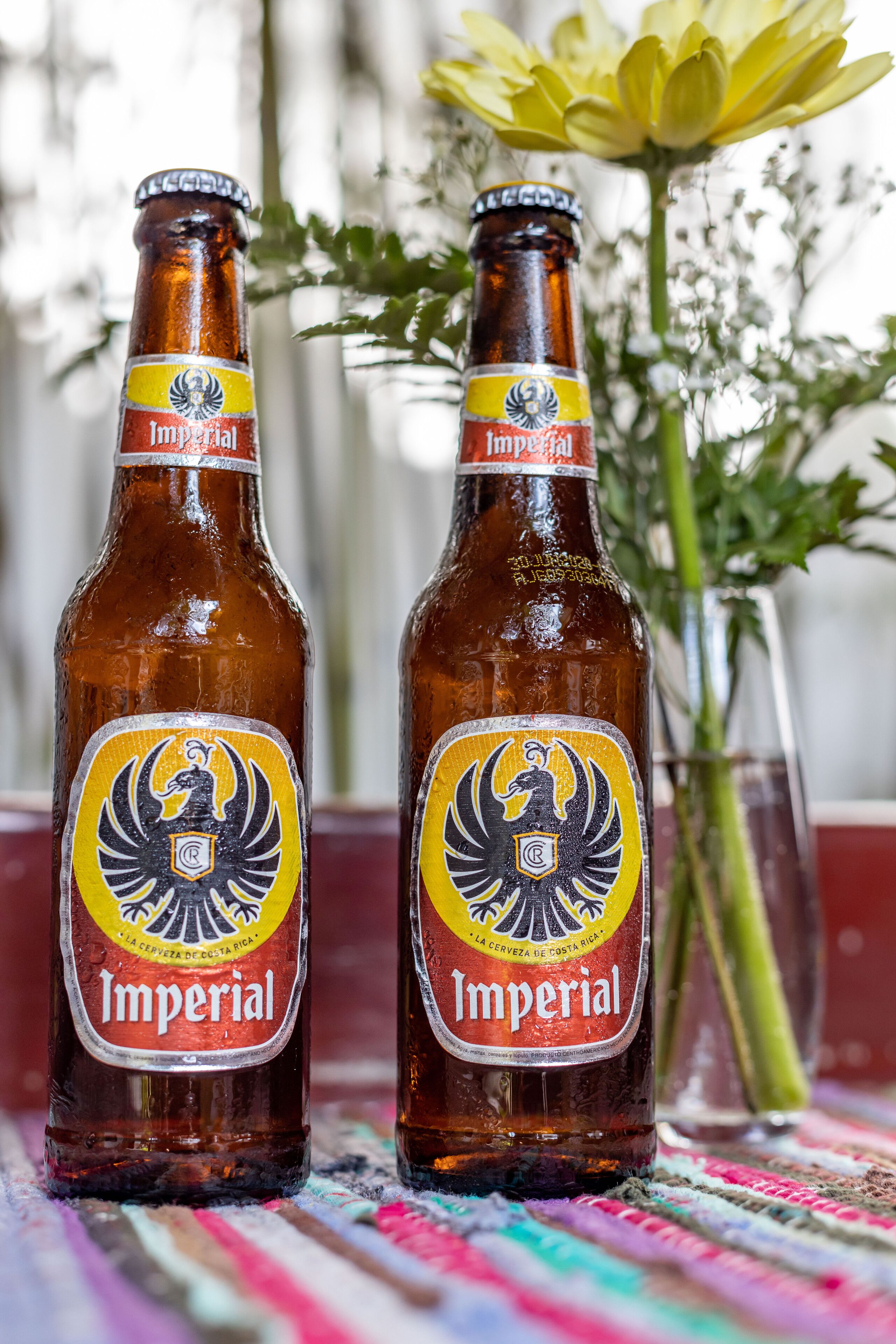 Two bottles of Imperial beer with a yellow label and a black eagle emblem, placed on a colorful woven surface.