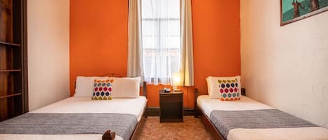 Una habitación con dos camas, una ventana con cortinas, una mesita y un cuadro en la pared.