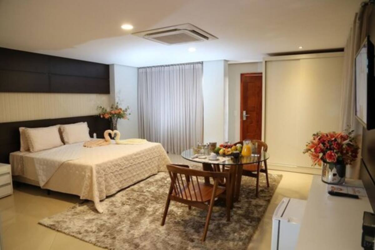 Habitación de hotel con cama, una mesa de comedor con sillas, refrigerador y ventana con cortinas.