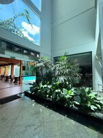 Un espacio interior moderno con pared de vidrio, zona de estar con mesas y sillas, y diversas plantas de interior.