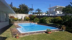 Piscina retangular com borda revestida de azulejos, rodeada por um deck de pedra e área gramada.