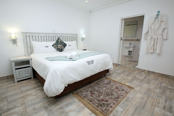 A neatly arranged bedroom with a bed, bedside tables, a nightstand, a bathroom visible through an open door, and a robe hanging on the wall.