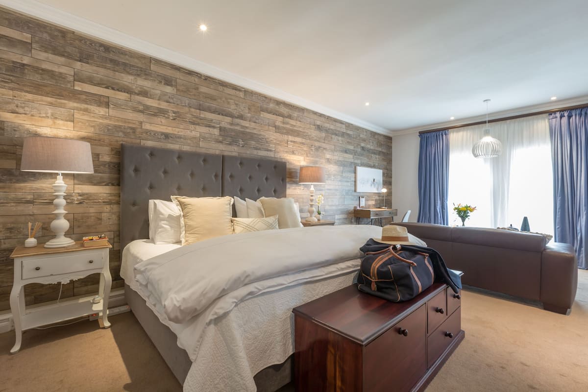 A modern bedroom with a wooden accent wall, a tufted headboard, a large bed, bedside tables with lamps, a dresser, and a view of the outdoors through a window with curtains.