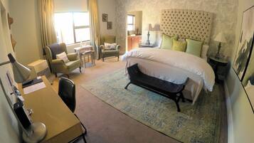 Una habitación de hotel con una cama grande, un área de descanso con un sofá y sillones, un escritorio con lámpara y una mesita con lámpara.