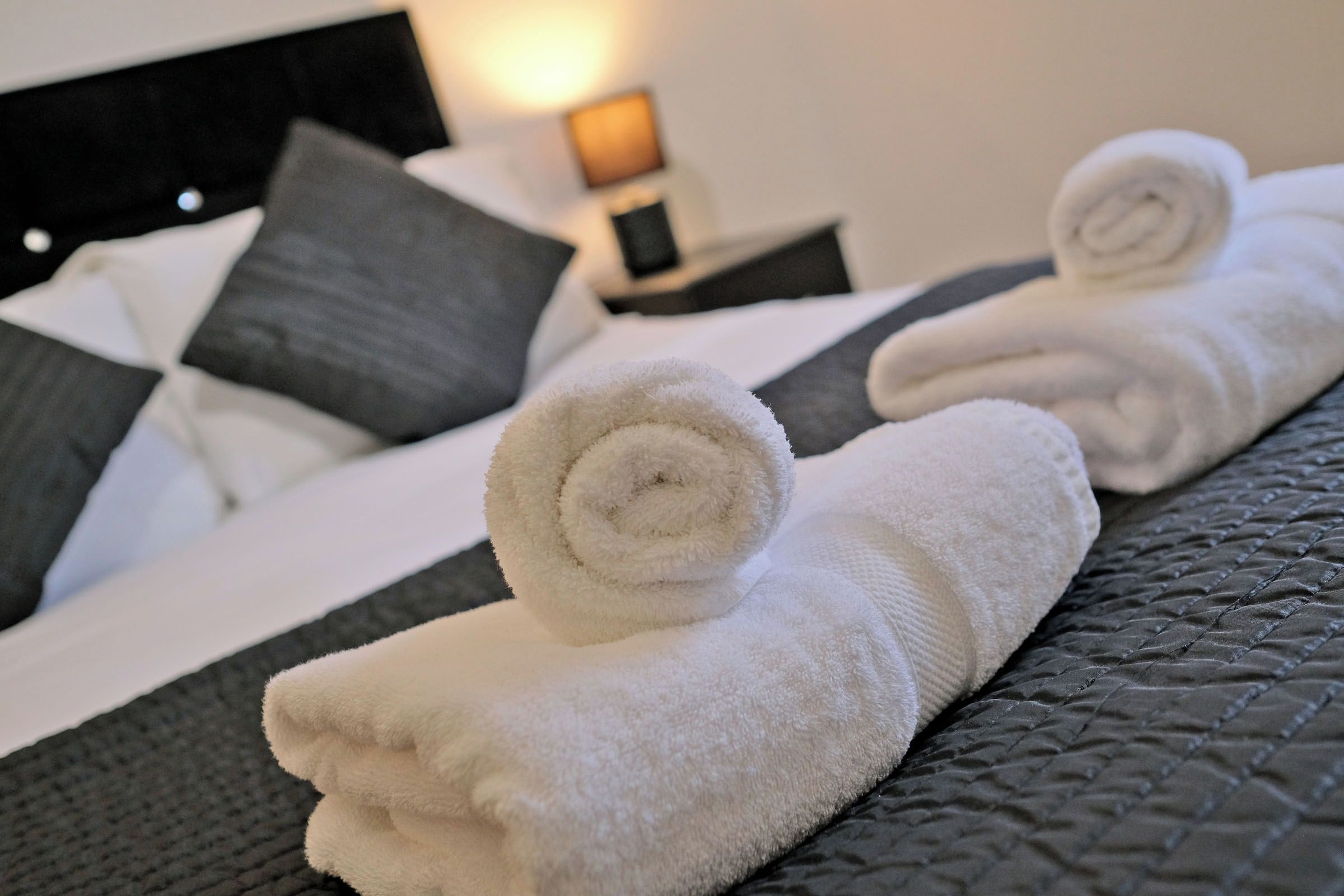 A neatly made bed with a white duvet, grey pillows, and a rolled white towel on the bed.