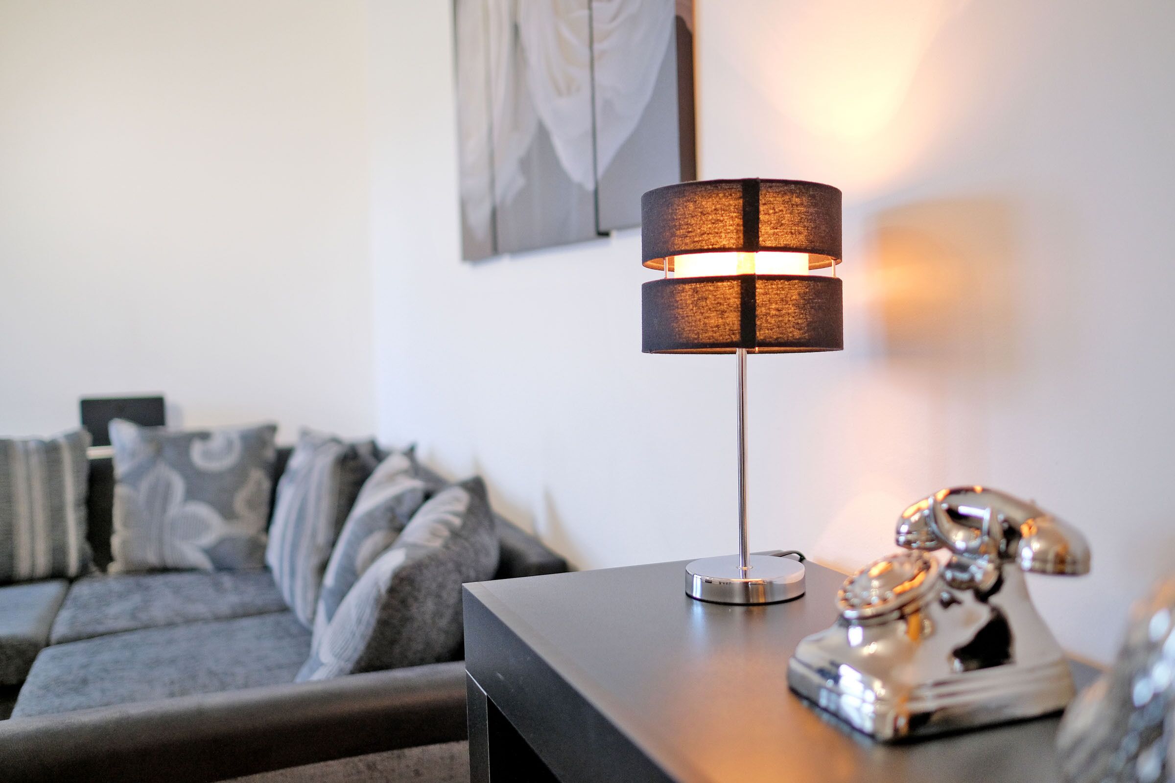 A modern living room with a sleek black table lamp, a vintage rotary phone, and a gray sofa with patterned cushions.