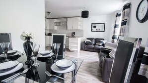 A modern living room with a glass dining table set for four, a sofa, and a kitchen in the background. - Am Pm Cults Townhouses (Aberdeen)