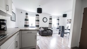 A modern kitchen with white cabinets, a wooden countertop, and a built-in dishwasher. There is a grey sofa in the living area and a dining table with chairs. - Am Pm Cults Townhouses (Aberdeen)