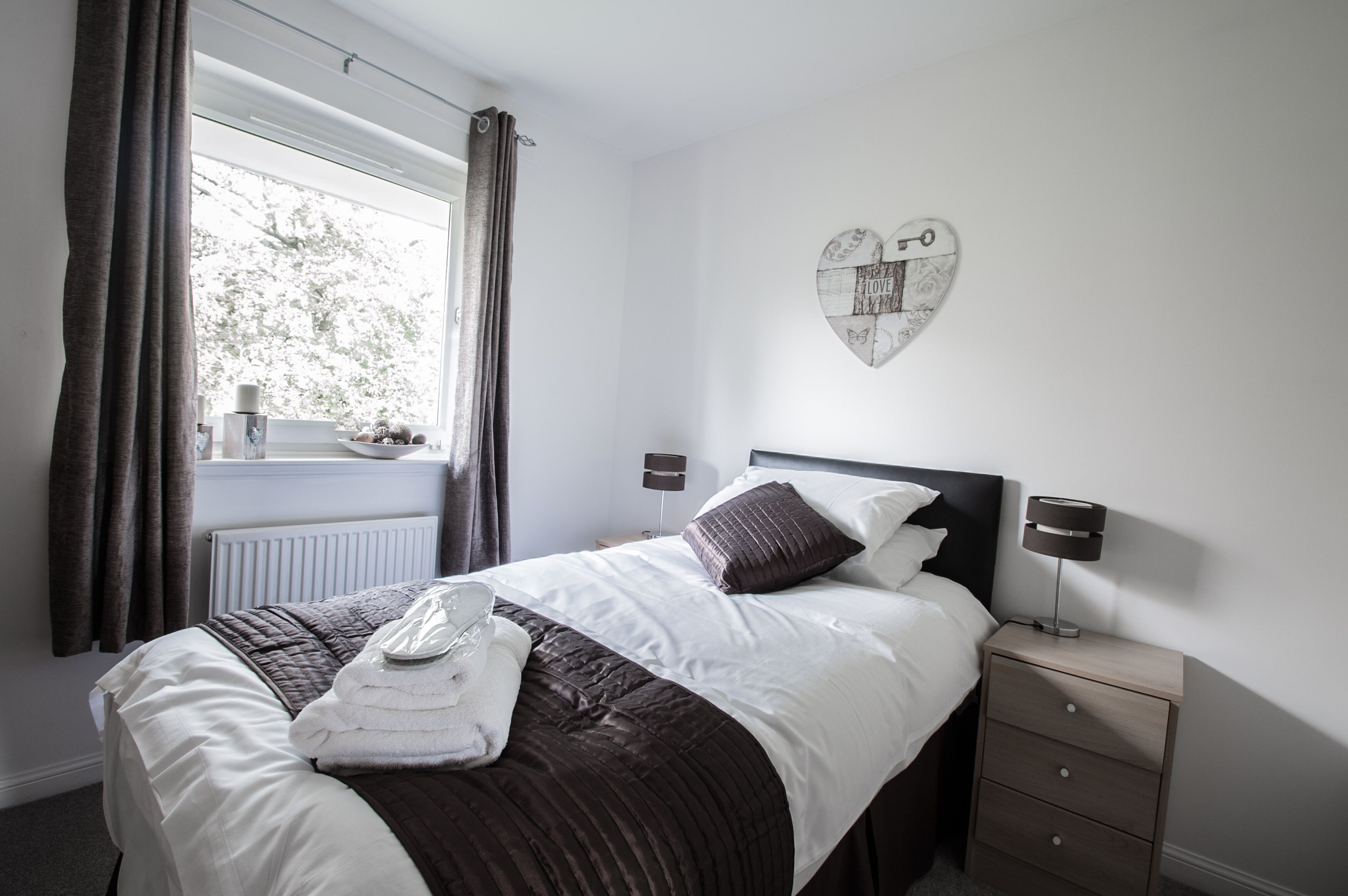 A neatly made bed with a heart-shaped decoration on the wall, a bedside table with a lamp, and a window with curtains.