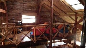 Una cabaña rústica de madera con cama en un altillo, una mesa de comedor y un ventanal.