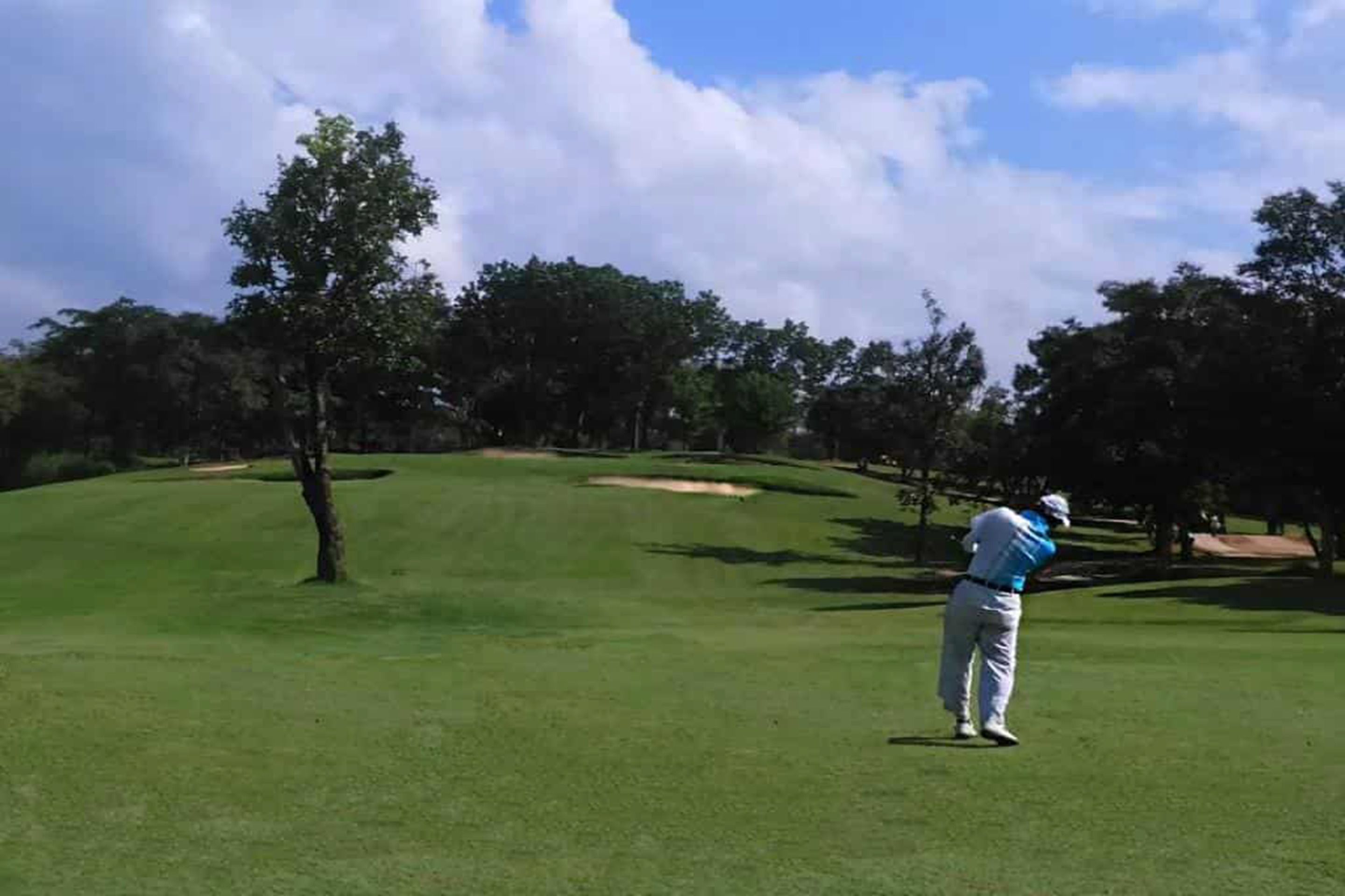 Un campo de golf con cielo despejado, árboles y un golfista en movimiento.