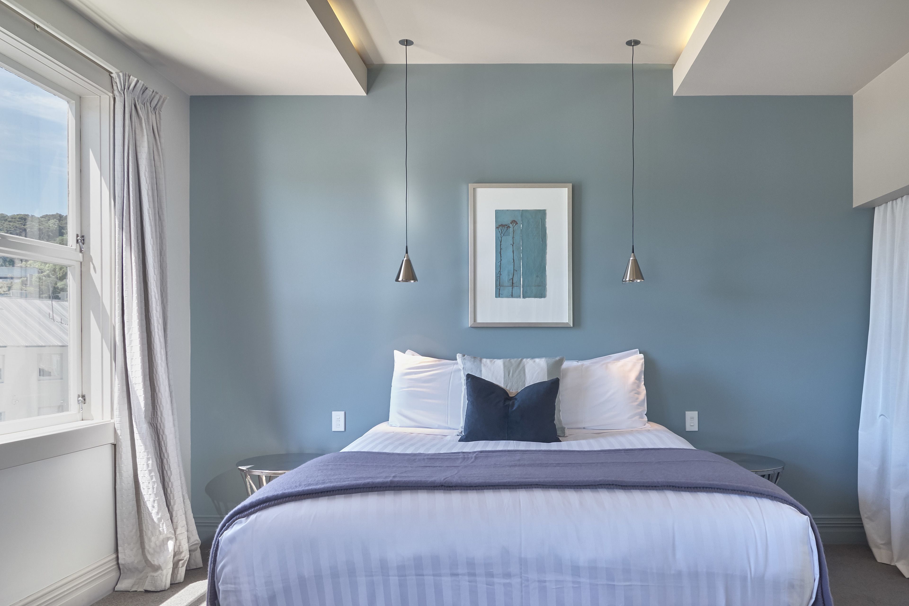 A bedroom with a bed, two bedside tables, a framed artwork, and a window with curtains.