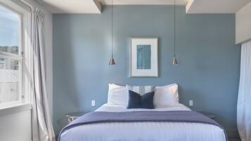 A bedroom with a bed, two bedside tables, a framed artwork, and a window with curtains.