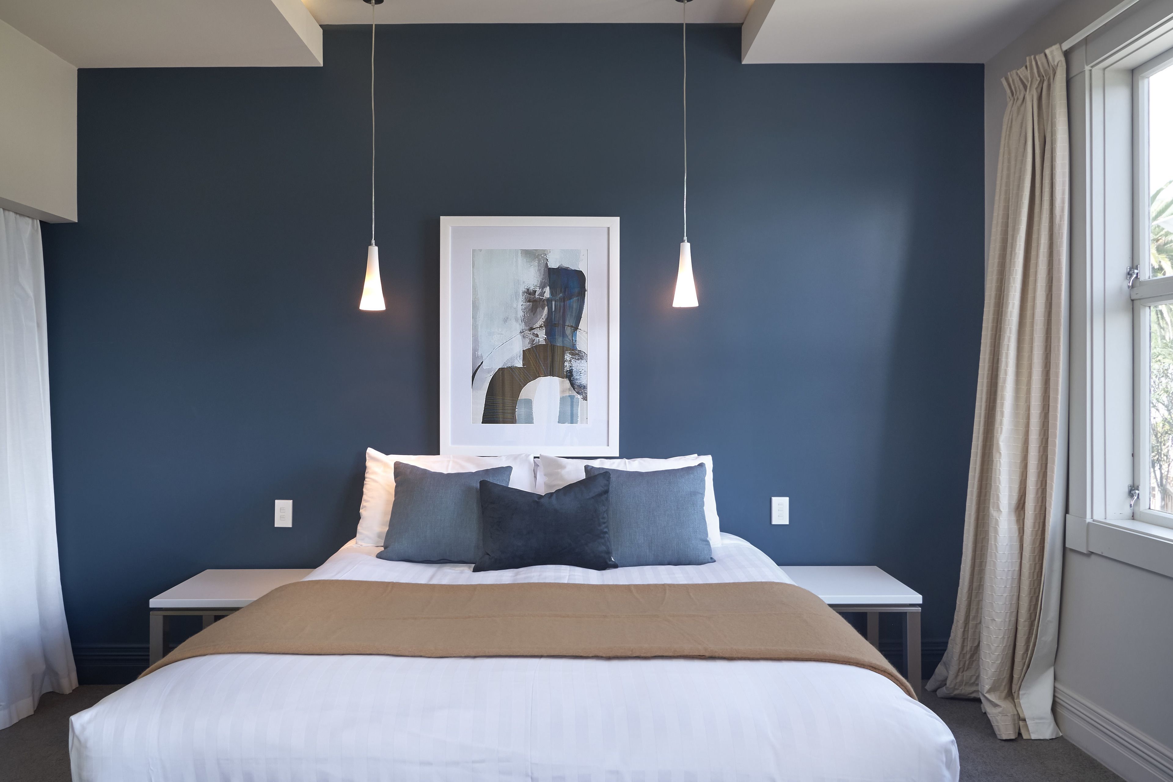 A bedroom with a bed, bedside tables, a framed abstract painting, and a window with curtains.