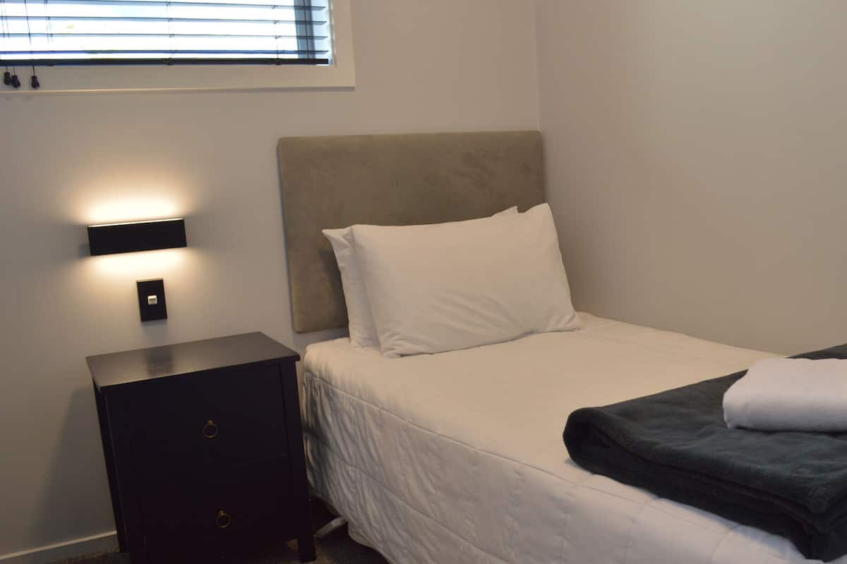 Ein ordentlich bezogenes Bett mit Kopfteil, ein Nachttisch mit Lampe und ein Fenster mit Jalousien.